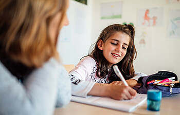 Ein Mädchen sitzt lächelnd am Tisch und schreibt gerade etwas in ihr Schulheft. Eine junge Frau sitzt daneben, man sieht diese nur von hinten.