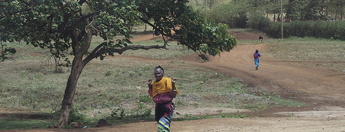 Eine afrikanische Frau geht einen Weg entlang rechts von ihr steht ein Baum.