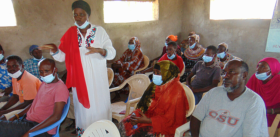Frauen und Männer der Gemeinde Loglogo sitzen in Stühlen in einer Gemeindebehausung und hören der Projektverantwortlichen zu.