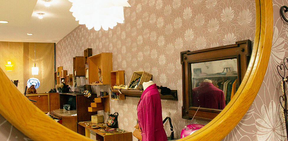 In einem Spiegel des carla+ sieht man eine Schaufensterpuppe mit einem pinken Oberteil und die gefüllten Regale des Geschäfts.
