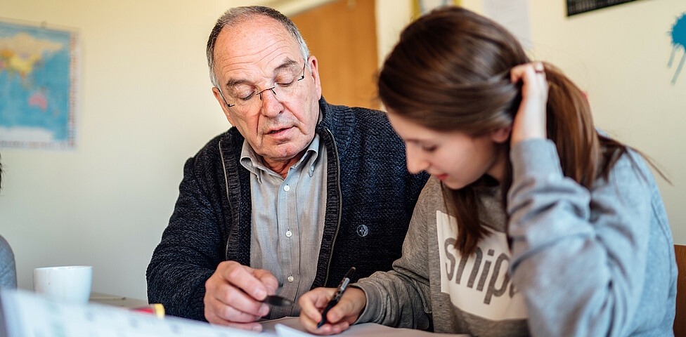 Ein älterer Herr mit Strickjacke sitzt bei einem jungen Mädchen am Tisch und erklärt ihr gerade eine Aufgabe, das Mädchen sieht konzentriert in ihr Heft.