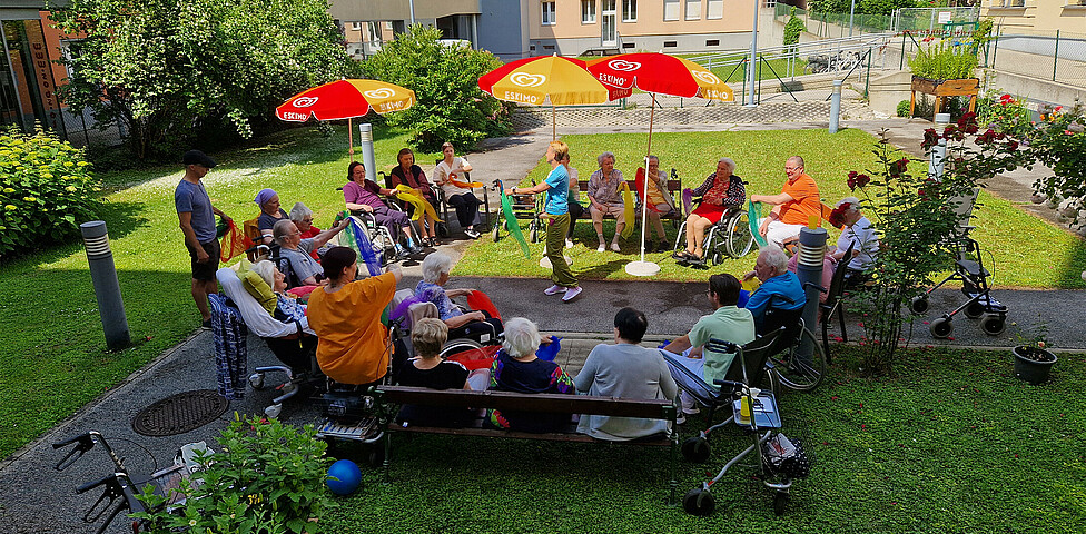 Eine Gruppe von Bewohner*innen sitzt in einem Kreis im Garten und mimt die Bewegungen mit den Tüchern der Betreuerin in der Mitte nach.