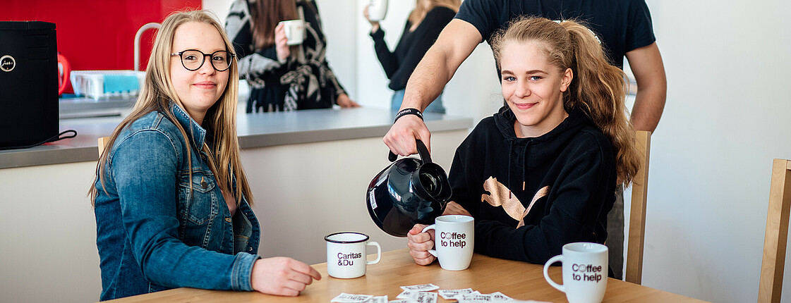 Zwei junge Frauen sitzen zu einem Coffee to help am Esstisch und ein junger Mann schenkt ihnen Kaffee ein. Im Hintergrund unterhalten sich zwei weitere Mädchen mit einer Tasse Kaffee in der Hand.