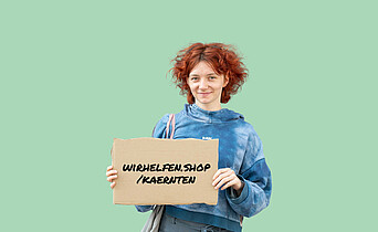Eine junge, rothaarige Frau hält ein Schild in der Hand auf dem wirhelfen.shop/kaernten geschrieben steht.