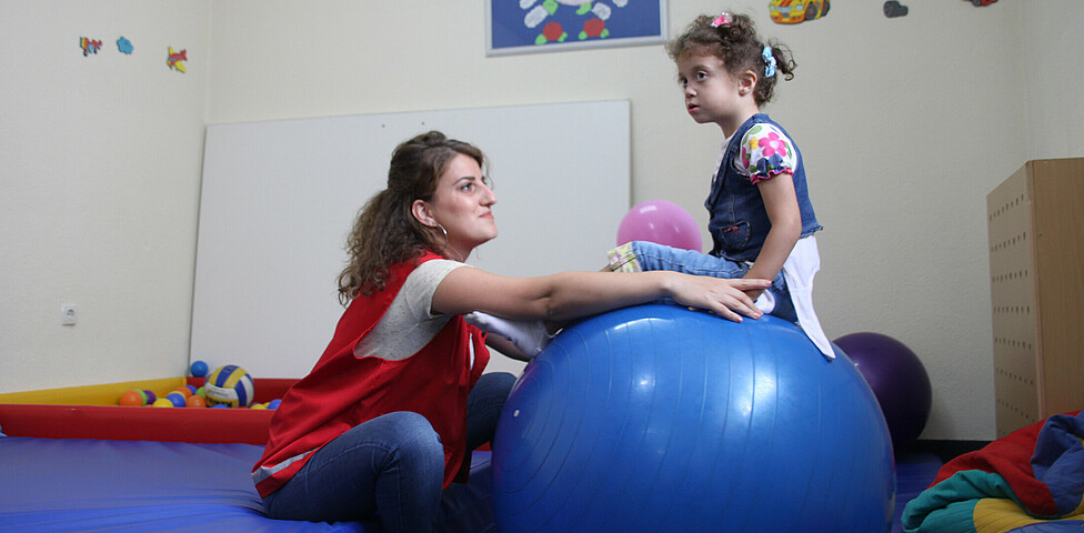 Eine Betreuerin hilft einem Mädchen mit dem Gleichgewicht, während es auf einem Gymnastikball sitzt.