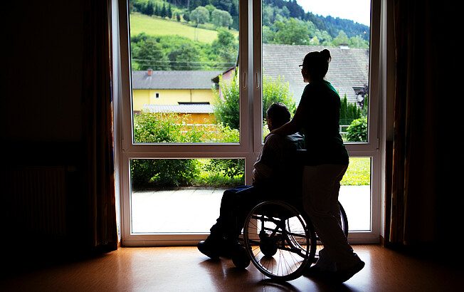 Eine Frau steht mit einem Menschen im Rollstuhl, die nur als Schatten gesehen werden, weil sie vor einem beleuchtenden Fenster stehen.