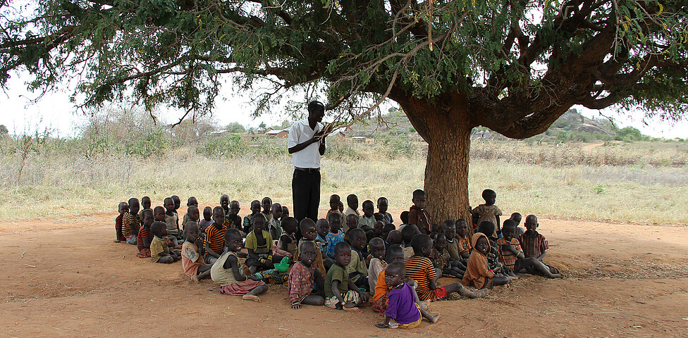 Im Schatten eines Baumes in Uganda werden viele Kinder von einem Lehrer unterrichtet.