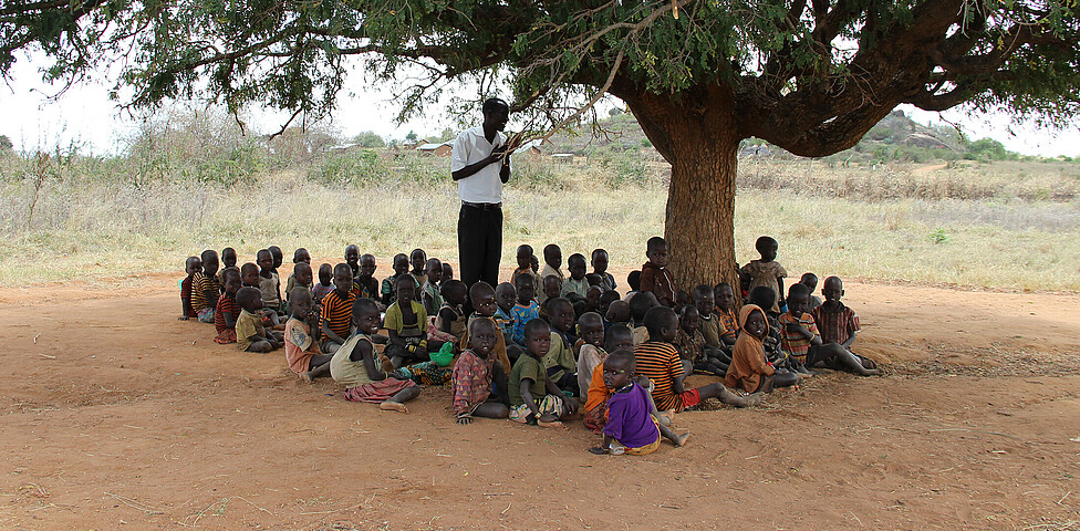 Im Schatten eines Baumes in Uganda werden viele Kinder von einem Lehrer unterrichtet.
