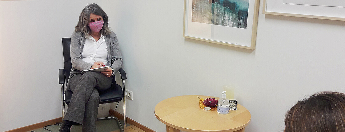 Eine Beraterin mit Mundschutz sitzt den Coronaabstand einhaltend vor einer Hilfesuchenden. Auf dem Tisch an der Wand steht eine Flasche Desinfektionsmittel und Taschentücher bereit.