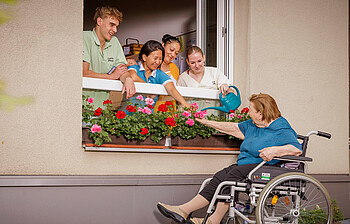 Vier Pflegekräfte lehnen sich aus dem Fenster, eine Bewohnerin sitzt im Rollstuhl und gießt die Blumen die auf der Fensterbank stehen.