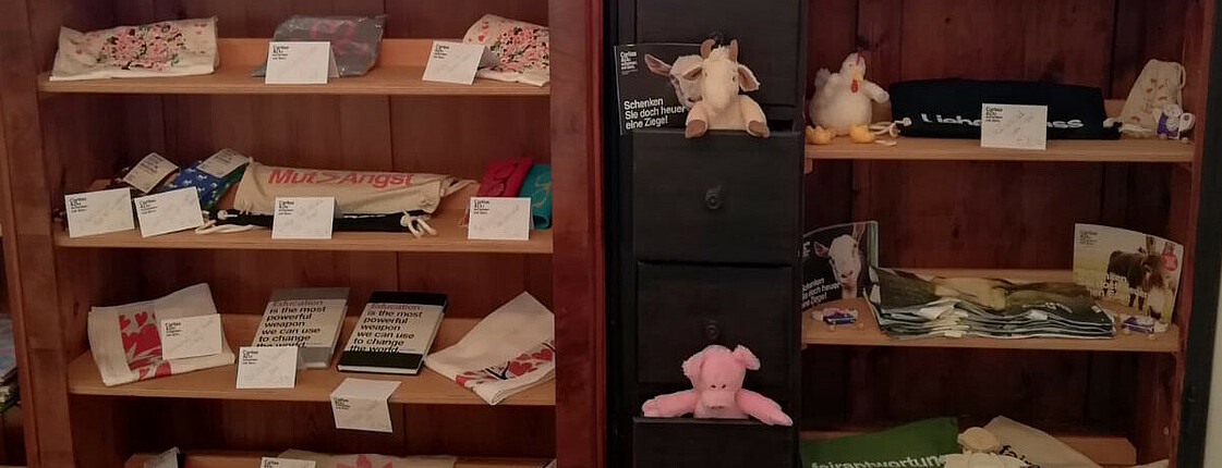 Zwei dunkle Regale gefüllt mit Produkten aus dem Online-Shop der Caritas Schenken mit Sinn. Man sieht T-Shirts, Taschen, Stofftiere und Socken.