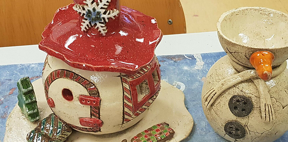 Rechts ein Gefäß, dass wie ein Schneemann geformt ist aus Ton, daneben ein Häuschen mit rotem Dach und Schneeflocke mit Schlitten vor dem runden Häuschen.