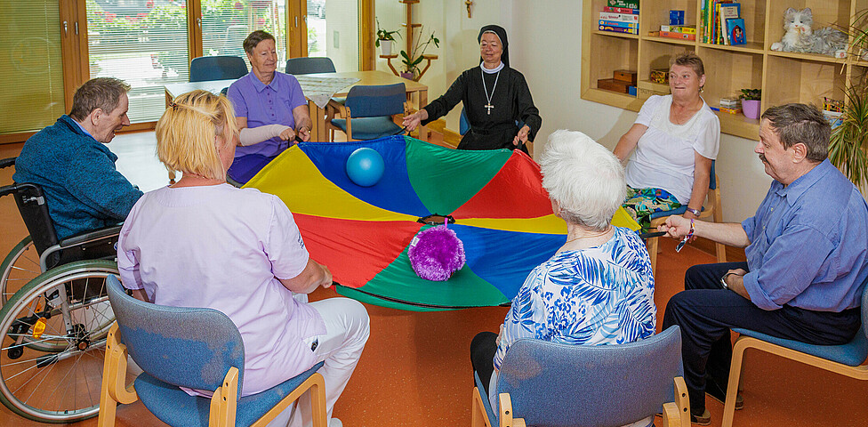 Eine Gruppe von Bewohner*innen sitzt im Kreis mit einem großen Tuch und sie befördern einen Ball nach recht und links.