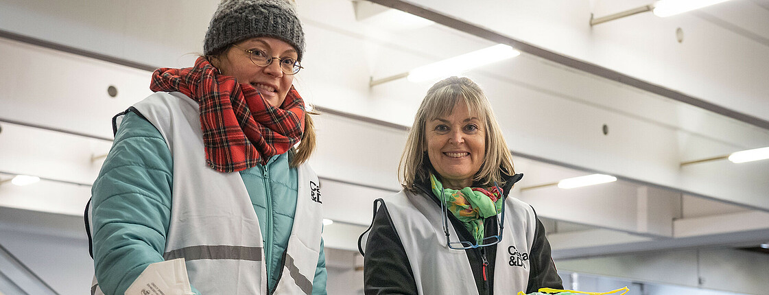Zwei Damen in Caritas-Warnwesten sortieren Kleidungsspenden in der Messehalle für die Ukrainehilfe.