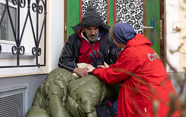 Ein obdachloser Mann sitzt dick eingepackt in Jacke und Schlafsack an einer Hausmauer. Eine Mitarbeiterin mit roter Caritas-Jacke hockt neben ihm und bietet Hilfe an.