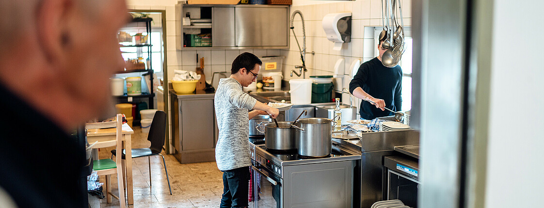 Ein Besucher unseres Eggerheims im Bild rechts und unscharf dargestellt, sieht in die Küche der Tagesstätte in der gerade zwei Zivildiener das Mittagessen vorbereiten.