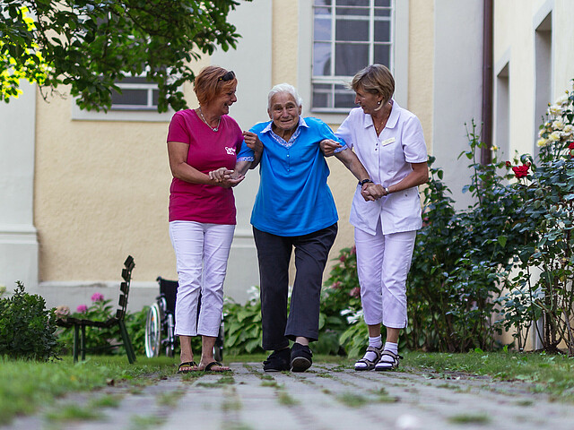 Zwei Pflegekräfte helfen einer älteren Dame, in dem sie diese an beiden Armen halten beim Spaziergang über einen gepflasterten Weg.