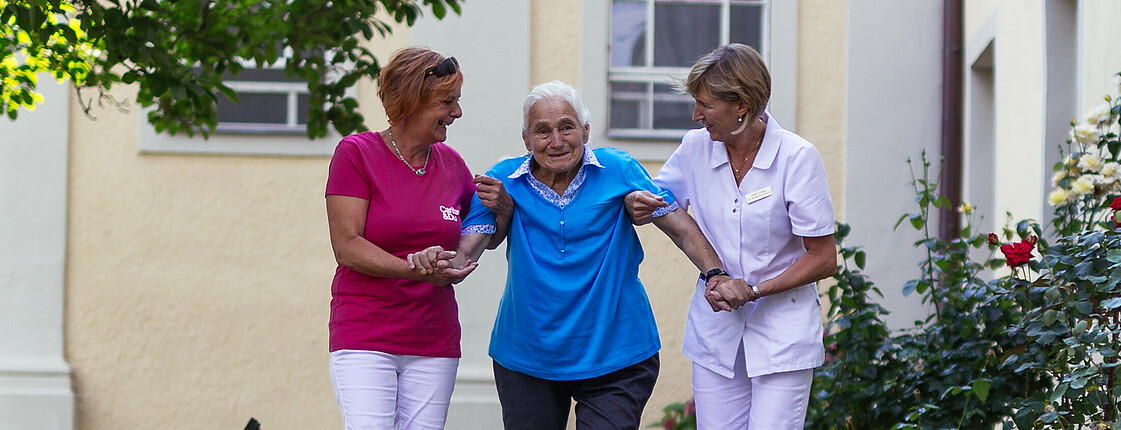 Zwei Pflegekräfte helfen einer älteren Dame, in dem sie diese an beiden Armen halten beim Spaziergang über einen gepflasterten Weg.