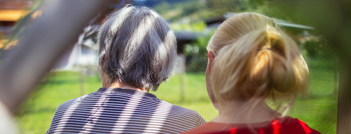 Eine junge Frau sitzt mit einer älteren Dame unter einer Laube mit den Rücken zugekehrt und sehen in den Garten.