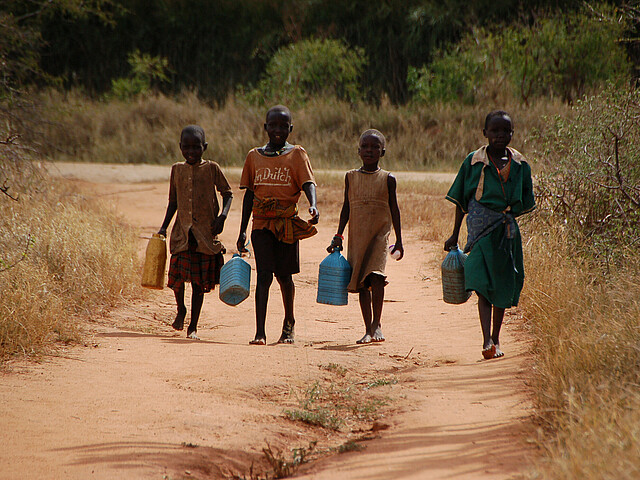 Vier Kinder sind auf einem Weg in Uganda unterwegs und tragen Wasserkanister.