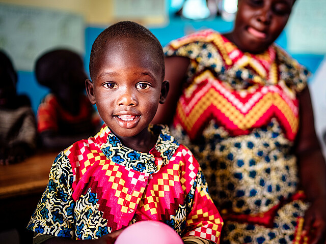 Ein afrikanisches Kind mit bunten Gewändern hat einen Luftballon in der Hand und lächelt in die Kamera, hinter ihm sitzt eine junge Frau ebenso in bunte Kleider gehüllt.
