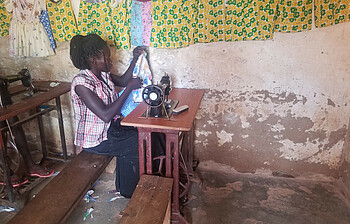 Eine junge Frau sitz an einer Nähmaschine und näht ein Kleidungsstück, als Teil Ausbildung.