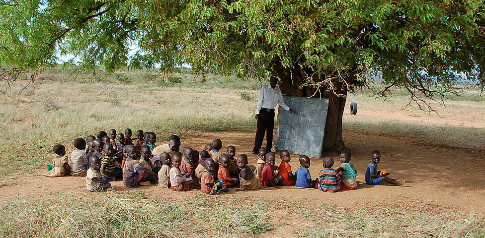 Eine Schar an Schüler*innen sitzt unter einem Baum und hat im Freien mit einem Lehrer der an der Tafel steht Unterricht.