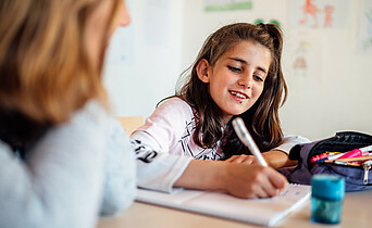 Ein junges Mädchen lächelt während sie gemeinsam mit einer freiwilligen Lernhelferin ihre Hausaufgaben macht.