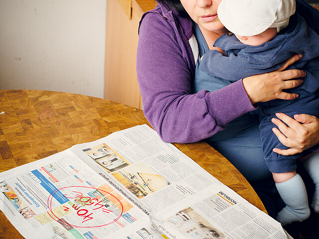Vor einer Frau, die ihr Kleinkind am Arm hat liegt eine Zeitung mit Wohnungsanzeigen.