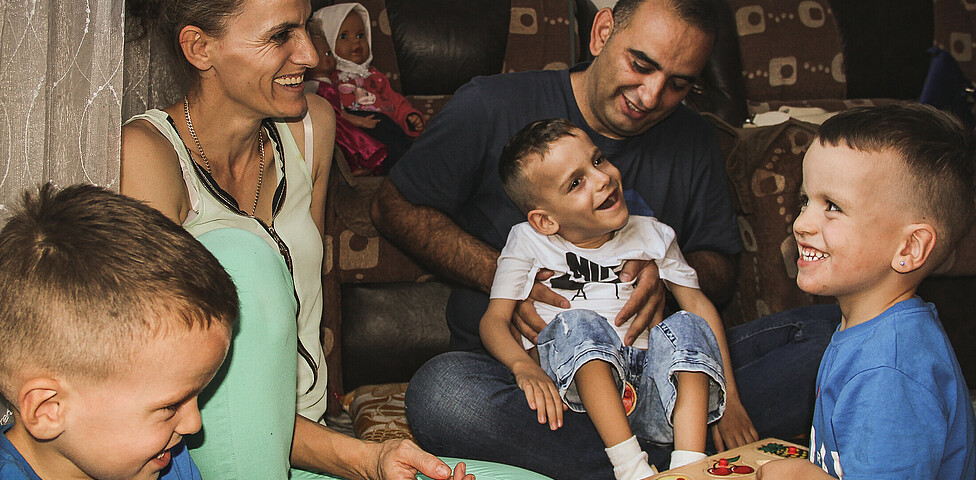 Doni ein Junge mit Behinderungen sitzt am Schoß seines Betreuers des Frühförderungszentrums und lacht gemeinsam mit seinen Zwillingsbrüdern und seiner Mutter.