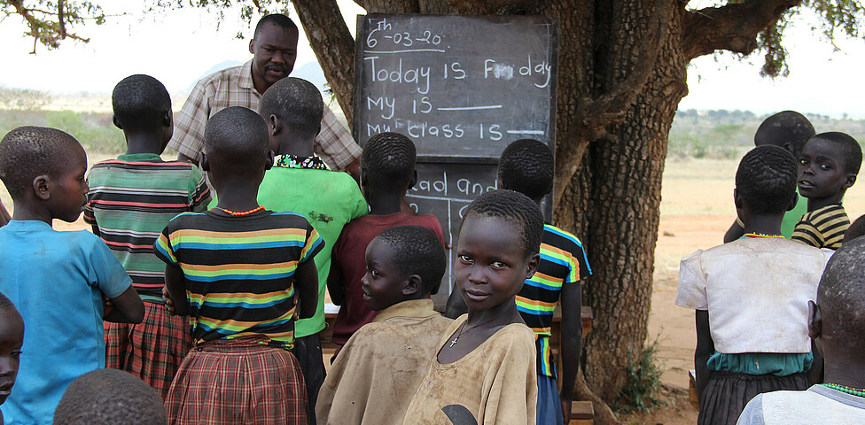An einem Baum ist eine Tafel angebracht, die afrikanischen Kinder stehen vor diesem und haben mit einem Lehrer Unterricht.