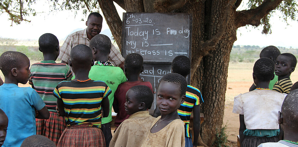 An einem Baum ist eine Tafel angebracht, die afrikanischen Kinder stehen vor diesem und haben mit einem Lehrer Unterricht.