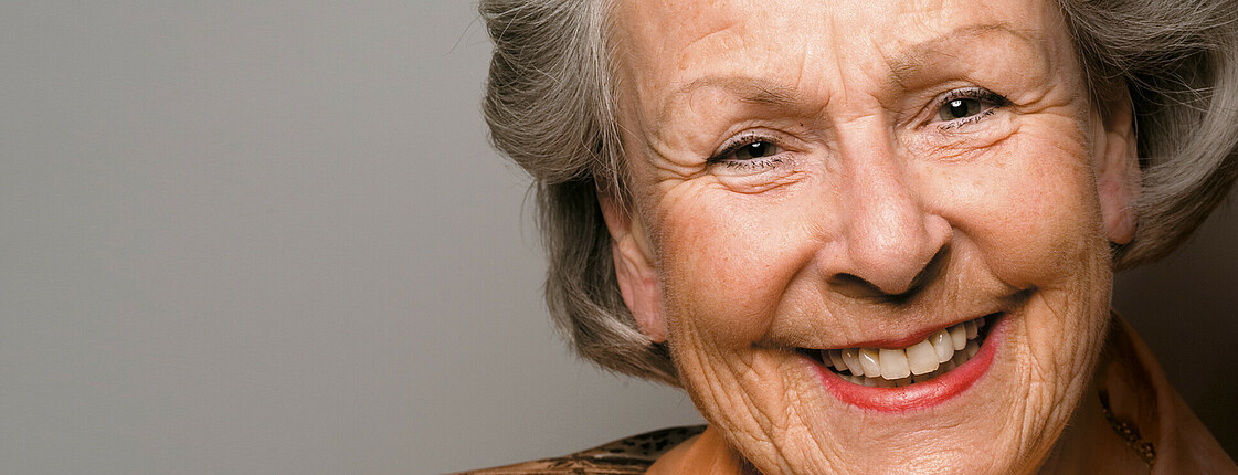 Eine ältere Dame, die schick hergemacht ist, lächelt in die Kamera.