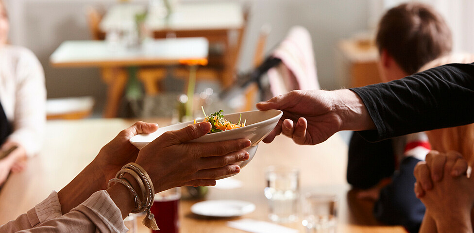 Eine Hand eines Kelleners überreicht einer Gästin einen Mittagssalat.