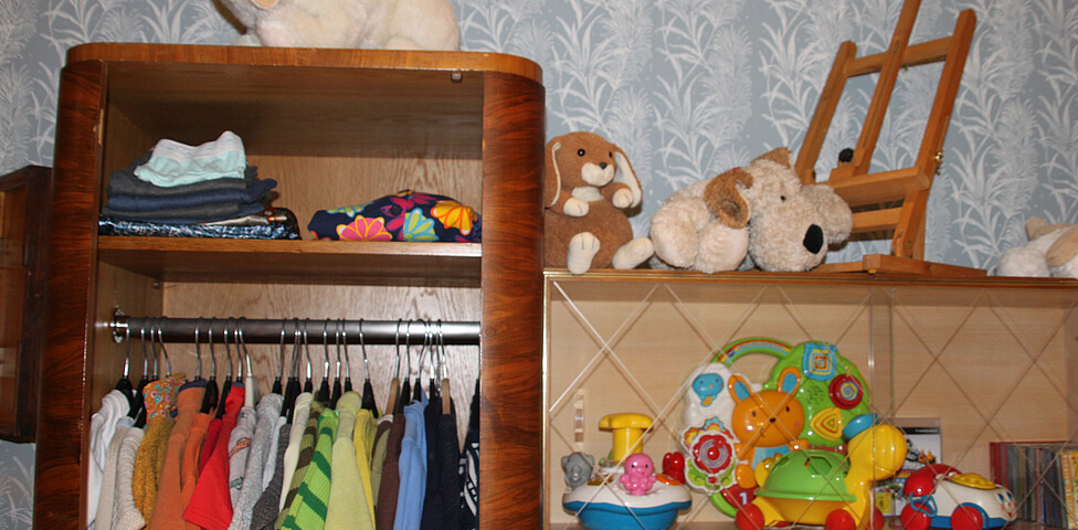 An der Wand ein Regal, links hängt Kinderbekleidung, rechts sind Spielzeuge an Regal.