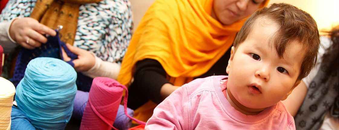 Zwei Damen mit Kopftuch stricken im Hintergrund während ein Kleinkind im Vordergrund am Tisch sitzt und in die Kamera sieht.