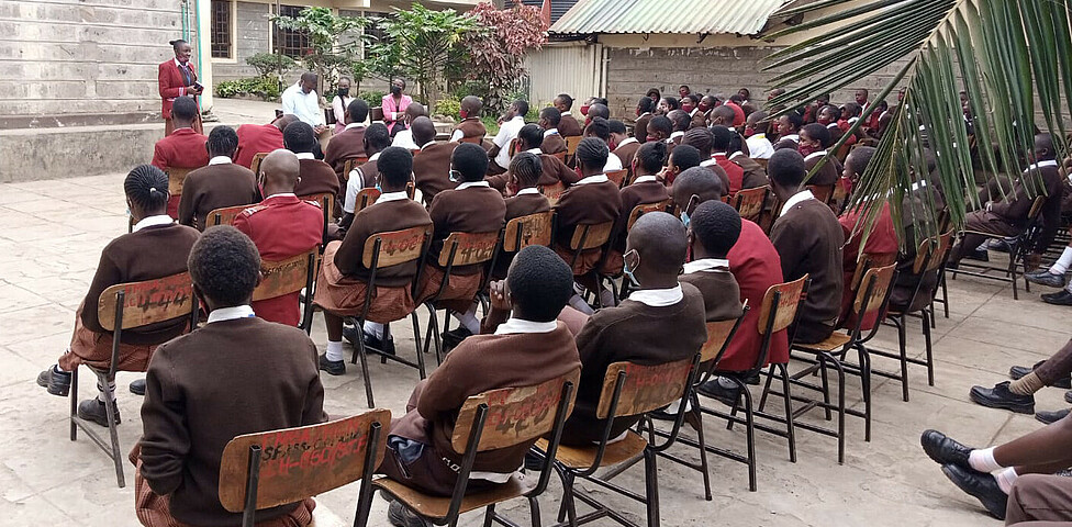 Schüler*innen sitzen im Innenhof der Schule und hören der Versammlung zu.