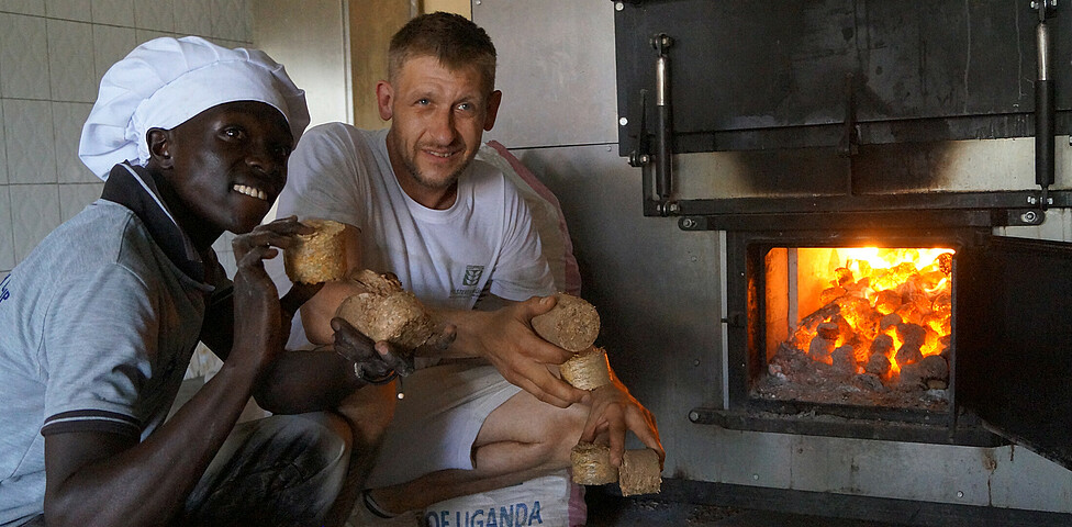 Ein Mitarbeiter der Bäckerei und der österreichisches Bäcker Josef knien vor dem großen Backofen und halten das Heizmaterial mit der Ofen beheizt wird in den Händen.