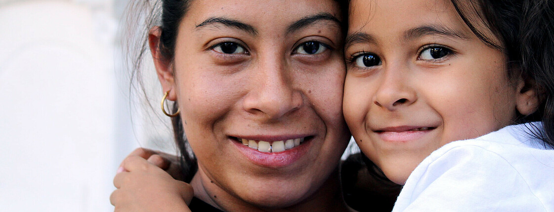 Eine Mutter hat ihre Tochter am Arm und beide lächeln in die Kamera.