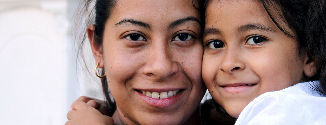 Eine Mutter hat ihre Tochter am Arm und beide lächeln in die Kamera.