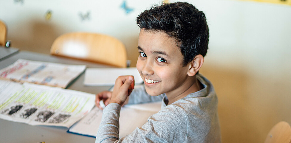 Ein Schüler sitzt an mit seinem Federpenal, Heft und einem Übungszettel am Tisch und lächelt in die Kamera.