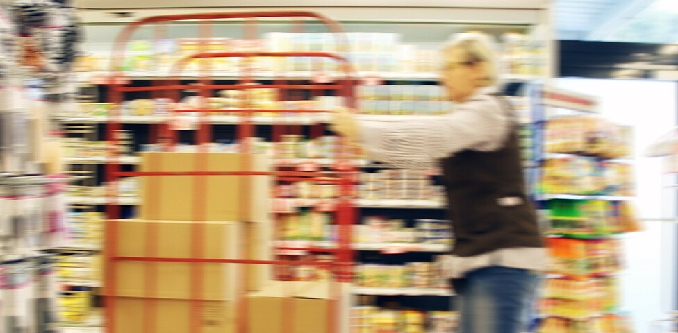 Eine SPAR Supermarkt Perspektive Handel-Mitarbeiterin fährt mit einem Rollwagen und neuen Waren durch den Gang des Supermarktes.