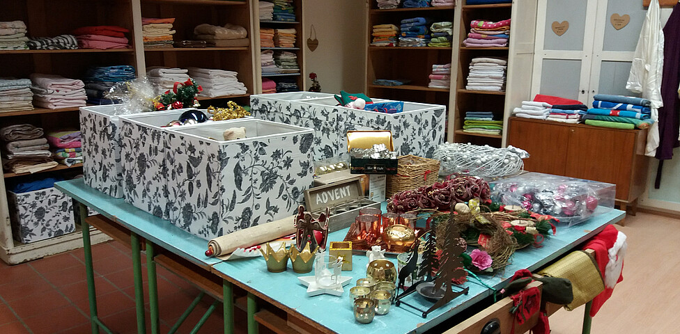 Auf einem großen Tisch liegen Kisten mit vielen Dekorationsartikeln für Weihnachten und an der Rückwand sind Regale voll mit Handtüchern.