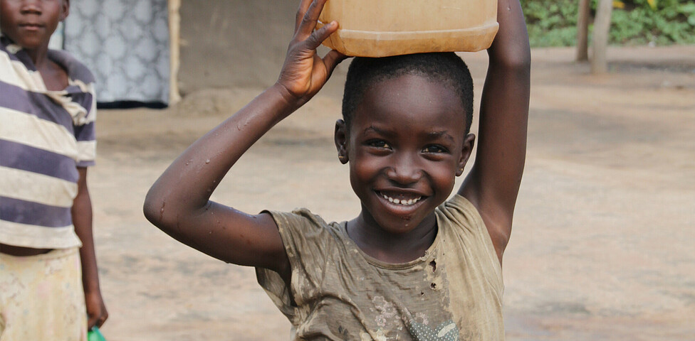 Ein Kind mit einem Wasserkanister am Kopf lächelt in die Kamera.