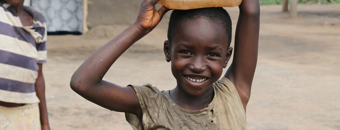 Ein Kind mit einem Wasserkanister am Kopf lächelt in die Kamera.