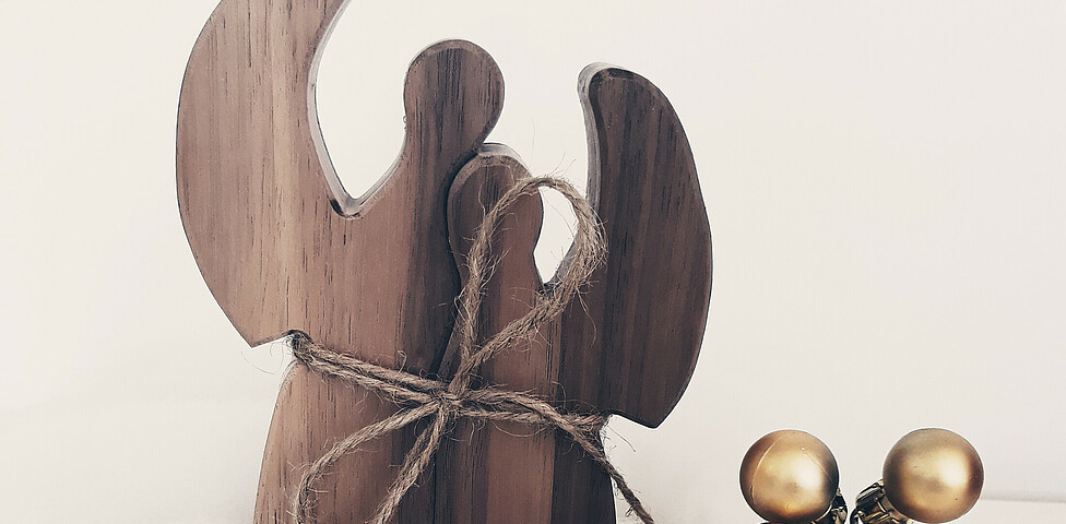 Zwei Engel aus Holz sind auf Watte und mit goldenen Kugeln verziert vor einem weißen Hintergrund drapiert.