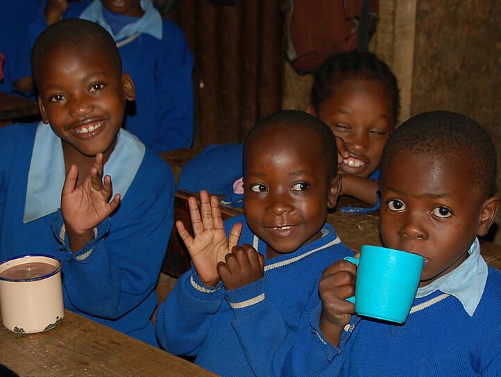 Afrikanische Kinder sitzend auf der Schulbank mit einem Trinkbecher in der Hand.